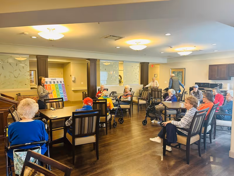 A group of elderly people seated around tables in a well-lit common room, participating in a game of Jeopardy led by a woman standing next to a large colorful Jeopardy board. The room has wooden flooring, comfortable chairs, and a kitchenette area in the background.