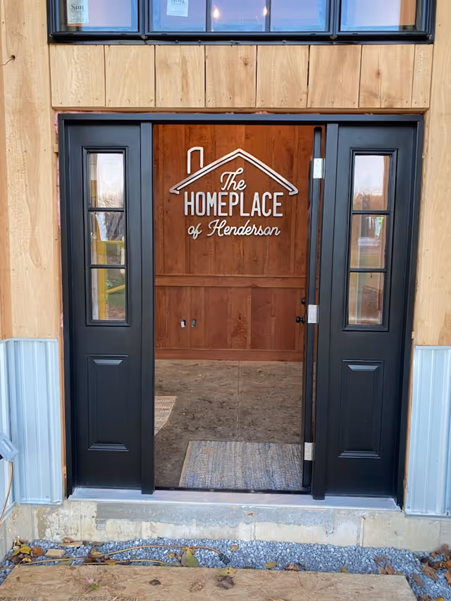Entrance doorway of a building under construction with black framed glass doors. Inside, a wooden wall displays a sign with a house roof graphic and the text 'The Homeplace of Henderson'.
