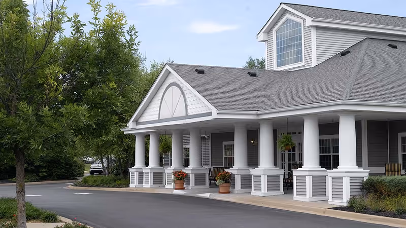 Exterior view of a senior living facility building with gray siding, white columns, and a covered entrance. There are potted plants with flowers near the entrance and trees along the driveway.