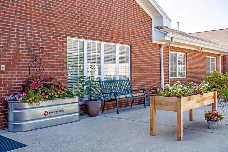 Outdoor patio area at Glen Ridge Health Campus with a brick exterior wall, two large windows with white blinds, a green metal bench with floral cushions, a metal planter filled with colorful flowers, a wooden raised garden bed with flowers, and a potted plant on the concrete ground.