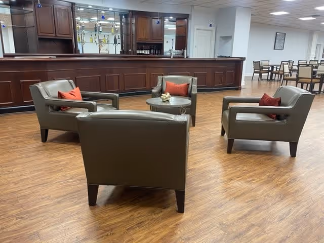 A spacious interior room with four gray leather armchairs arranged around a small round table with a decorative item on it. Each chair has a red cushion. Behind the seating area is a long wooden bar with shelves holding bottles and glasses. In the background, there are several tables and chairs arranged for dining or socializing. The floor is wood, and the ceiling has recessed lighting.