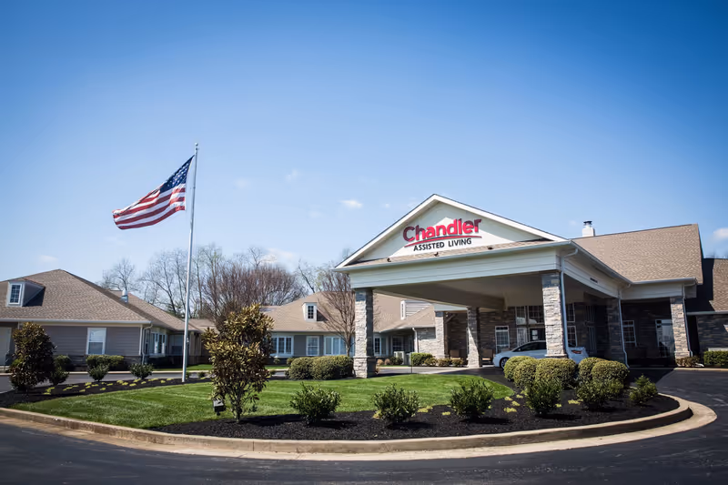 Exterior view of Chandler Park Assisted Living facility with a covered entrance, manicured bushes, green lawn, and an American flag flying on a flagpole under a clear blue sky.