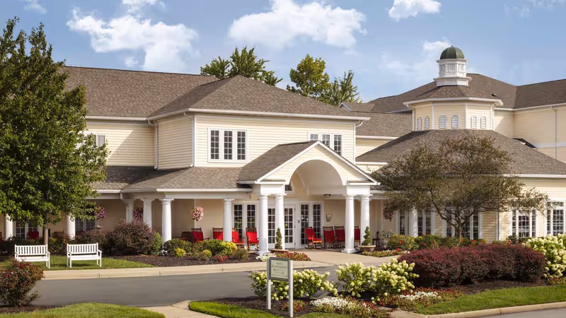 Front entrance of a two-story cream-colored senior living building with a covered portico, white columns, red rocking chairs on the porch, and landscaped gardens.