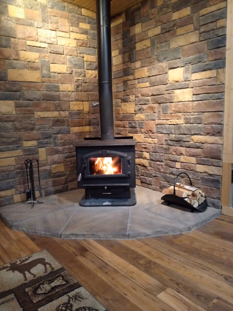 A stone-walled corner with a wood stove and fire burning, surrounded by a hearth and tools.