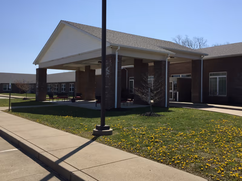 Exterior view of Breckinridge Place Retirement Community showing a covered entrance supported by brick columns, a sidewalk, a grassy area with dandelions, and a clear blue sky.