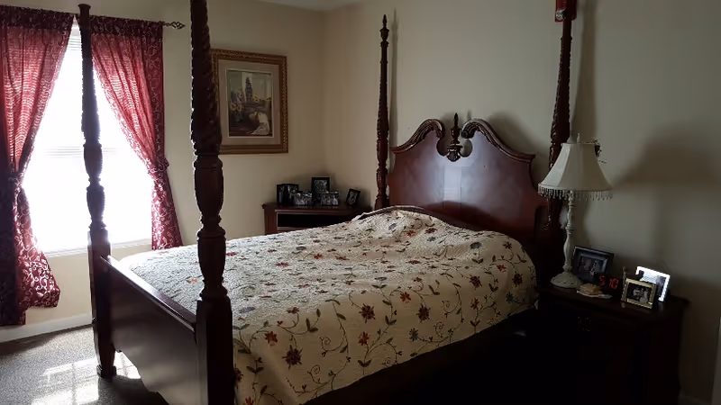 A bedroom with a large wooden four-poster bed covered with a floral quilt. There are two wooden nightstands on either side of the bed, one with a lamp and framed photos, and the other with several framed photos. A window with red curtains allows natural light into the room. A framed picture hangs on the wall above one nightstand.