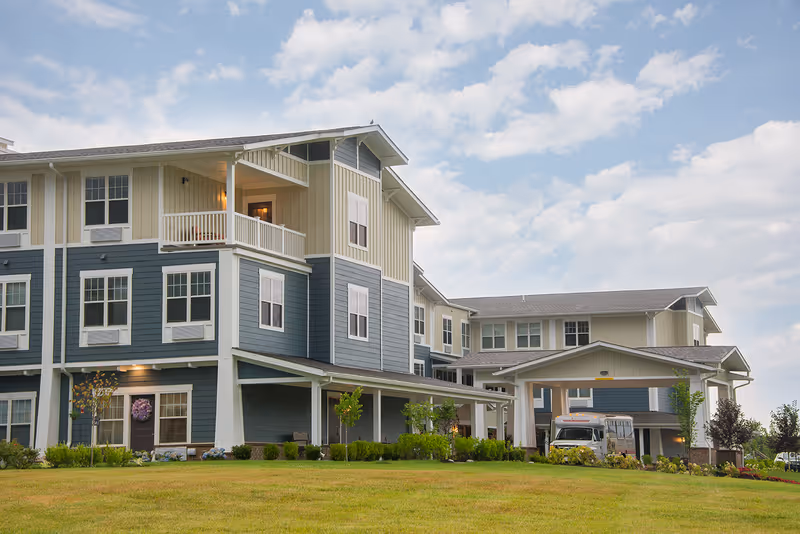 Exterior view of a multi-story senior living facility building with blue and beige siding, multiple windows, a covered entrance with a vehicle parked underneath, and a well-maintained lawn and landscaping in the foreground under a partly cloudy sky.