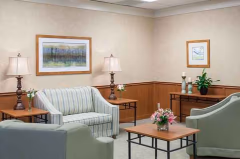 A cozy senior living lounge with striped sofa, green chairs, tables, and wall art.