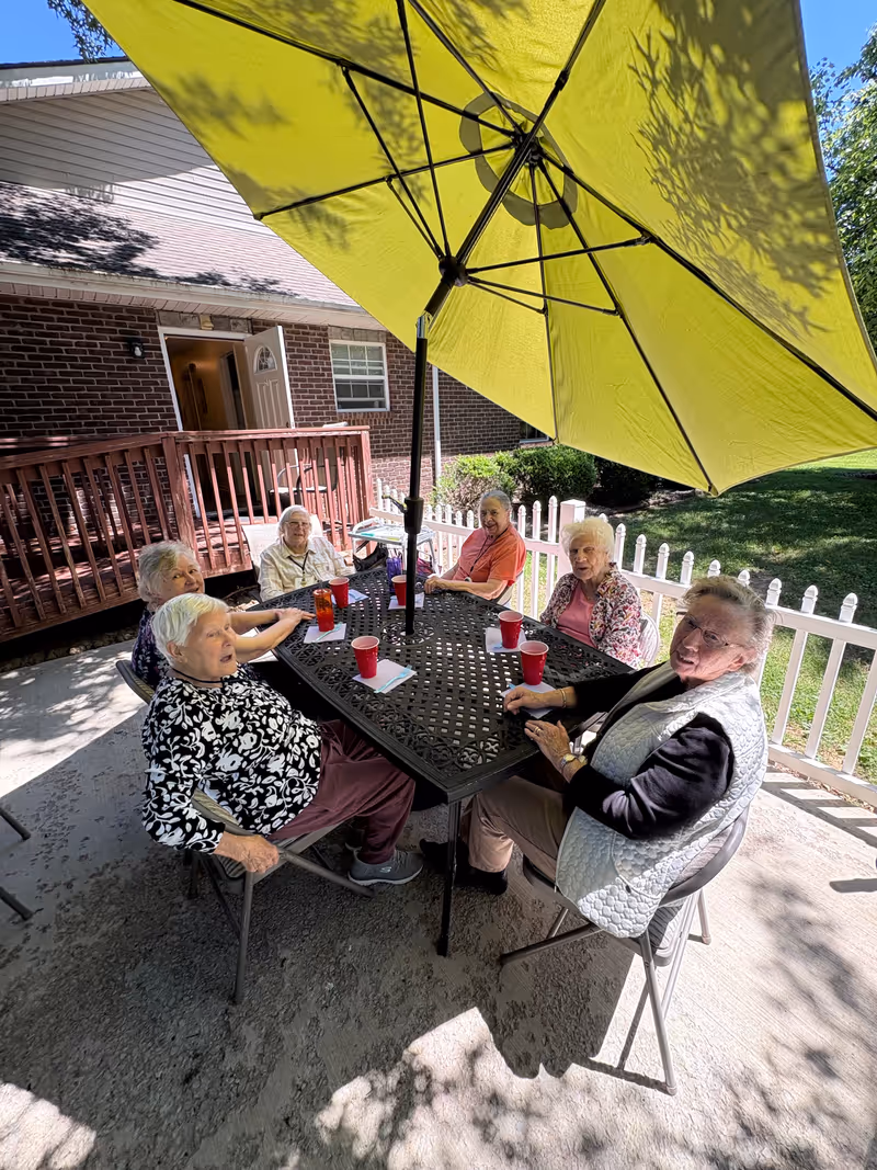 Six elderly residents sit around a metal table under a large yellow umbrella outdoors.