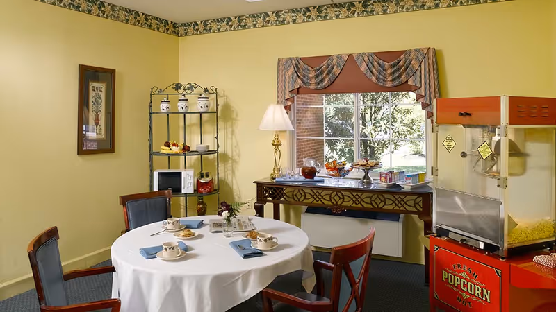A small dining area with a round table covered with a white tablecloth, set with four cups and saucers, blue napkins, and cookies. Behind the table is a window with a decorative valance, a table with snacks and drinks, a popcorn machine, and a metal shelving unit holding a microwave and decorative jars. The walls are painted yellow with a floral border near the ceiling.