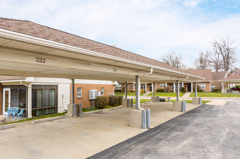 Covered parking spaces numbered 322 and 314 in front of a single-story brick and siding building with windows and a blue bench outside. The area is paved with a driveway and some grass and trees in the background under a partly cloudy sky.