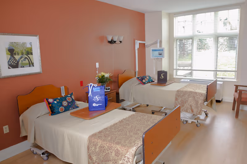 Two hospital-style beds in a bright senior living room with bedside tables, decorative pillows, and a large window.