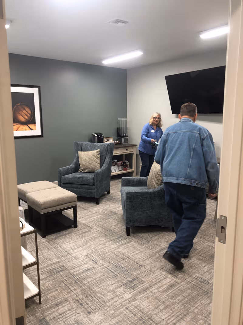 Indoor senior living lounge with blue chairs, a coffee station, and two people.