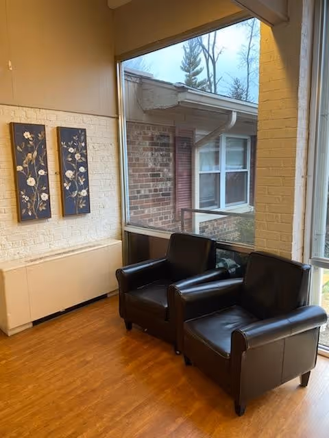 A cozy corner of a room with two black leather armchairs placed side by side near large windows. The windows offer a view of a brick building exterior with white-framed windows and a tree in the background. The interior walls are painted white with two decorative floral paintings hanging on one wall. The floor is wooden.