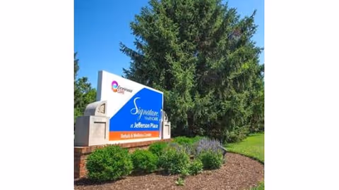 Entrance sign for Signature HealthCARE at Jefferson Place Rehab & Wellness Center surrounded by landscaped shrubs and a large evergreen under a blue sky.