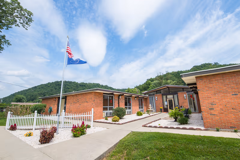 Exterior view of Prestonsburg Health Care Center showing a single-story brick building with a white picket fence, landscaped garden beds with flowers, a flagpole with the American flag and another flag, and a clear blue sky with some clouds and green hills in the background.