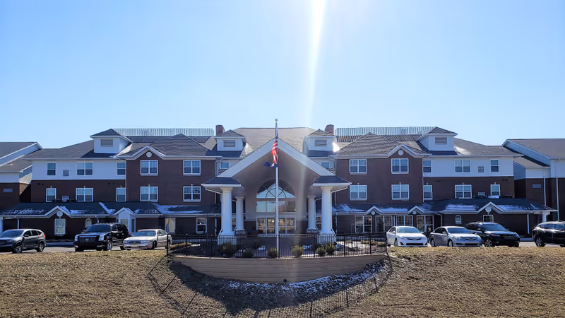 Front exterior view of Village Crossing Retirement Community building with a large covered entrance supported by white columns, multiple windows, and several cars parked in front under a clear blue sky.
