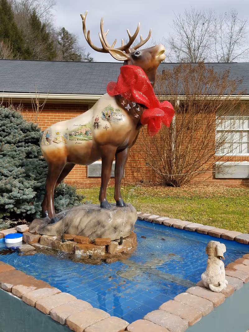 Statue of a deer with a red scarf atop a small fountain by a brick building