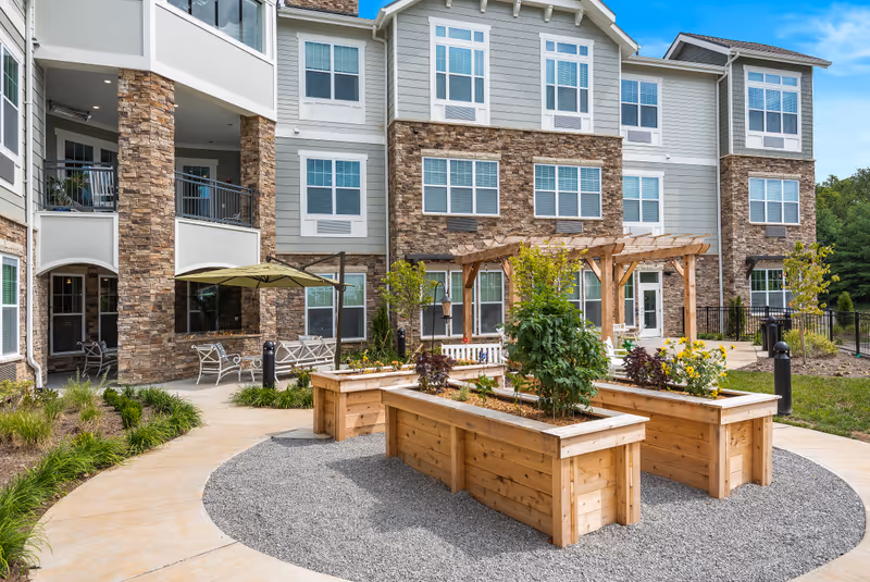 Outdoor garden area at The Ashton on Dorsey featuring raised wooden planter boxes with various plants and flowers, a wooden pergola, benches, and a stone and siding building in the background under a blue sky.