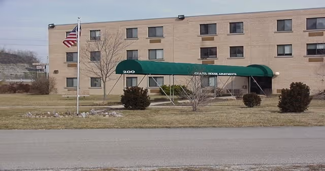 Exterior view of a three-story beige brick building with multiple windows. A green awning labeled 'CHAPEL HOUSE APARTMENTS' extends over the entrance, with the number 200 displayed on the side. An American flag is flying on a flagpole in front of the building, and there are some bushes and leafless trees on the lawn.
