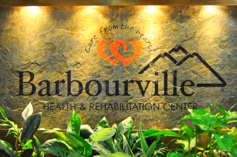 A stone lobby sign reading 'Barbourville Health & Rehabilitation Center' with a heart logo above and plants below.