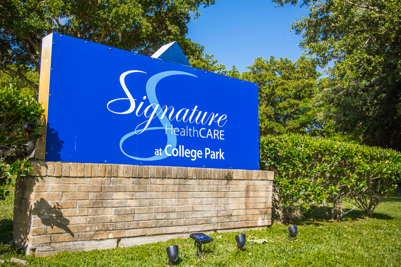 A large blue sign with white text reading 'Signature HealthCARE at College Park' mounted on a low brick wall, surrounded by green bushes and trees under a clear blue sky.