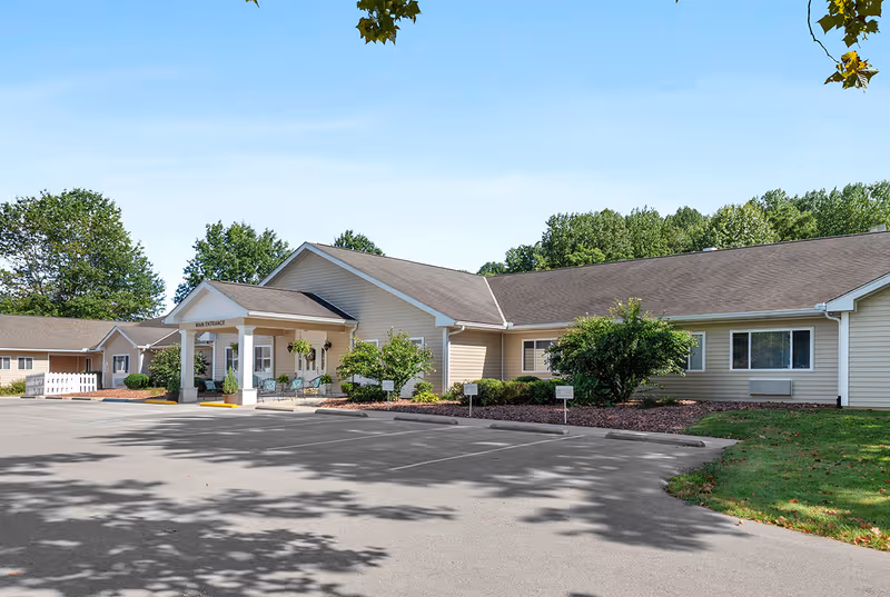 Exterior view of a single-story beige building with a covered main entrance, surrounded by greenery and trees under a clear blue sky. The parking lot in front is empty with visible parking spaces.
