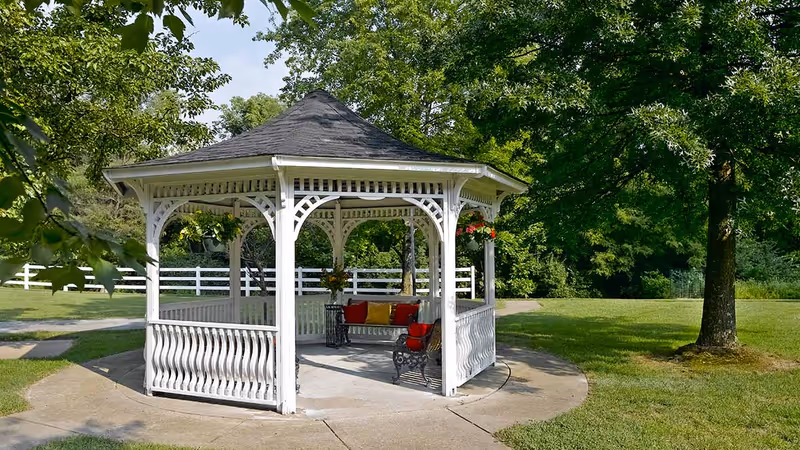 A white wooden gazebo with a shingled roof situated on a concrete circular pad in a grassy outdoor area. Inside the gazebo, there is a bench with red and yellow cushions and a small black metal table. The surrounding area features green trees, a white fence, and a clear sky.