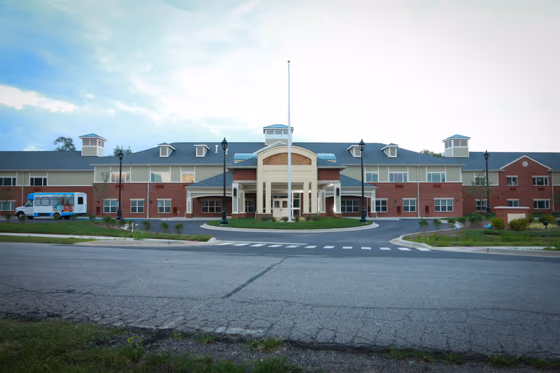 Front exterior view of a large two-story senior living facility building with a circular driveway and a flagpole in front. The building has a combination of brick and light-colored siding with multiple windows and small cupolas on the roof. A small shuttle bus is parked on the left side of the building.