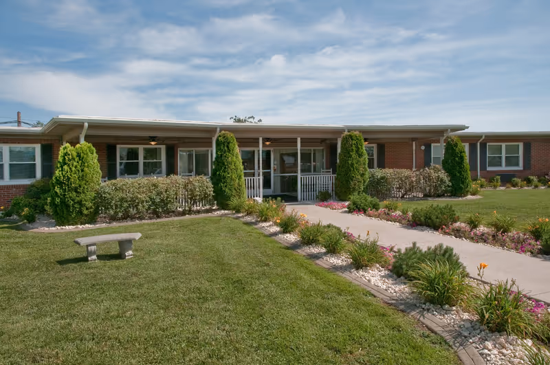 Front exterior view of a single-story brick building with a covered entrance, surrounded by neatly trimmed bushes, small trees, and a well-maintained lawn with a concrete walkway and flower beds on either side. A stone bench is placed on the grass near the walkway.