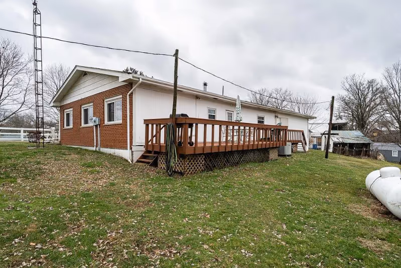 Exterior view of a single-story building with a brick and white siding facade, featuring a wooden deck with railings and steps leading down to a grassy yard. The sky is overcast and there are leafless trees in the background.