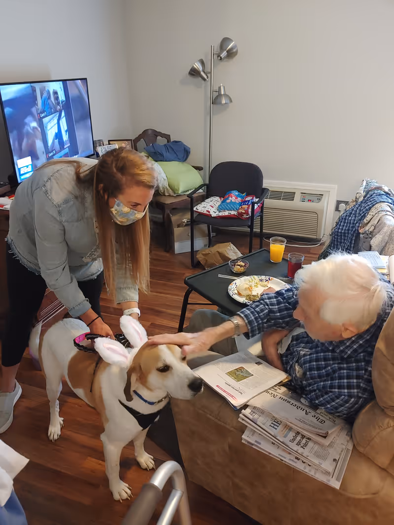 An elderly man sitting in a recliner chair inside a living room area, petting a dog wearing bunny ears. A woman wearing a face mask is holding the dog’s leash and smiling. The room has wooden floors, a TV in the background, a small table with food and drinks, and some chairs with cushions.
