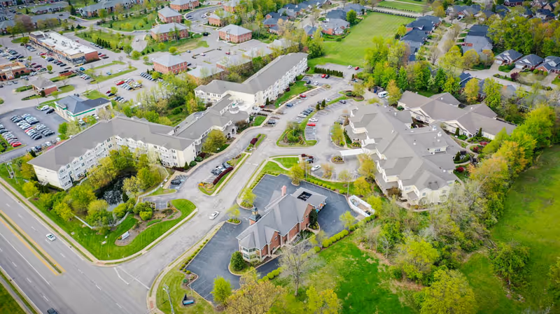 Aerial view of Arcadia Louisville senior living facility showing multiple large buildings surrounded by parking lots, roads, and green landscaped areas with trees and grass. Residential neighborhoods and other buildings are visible in the background.