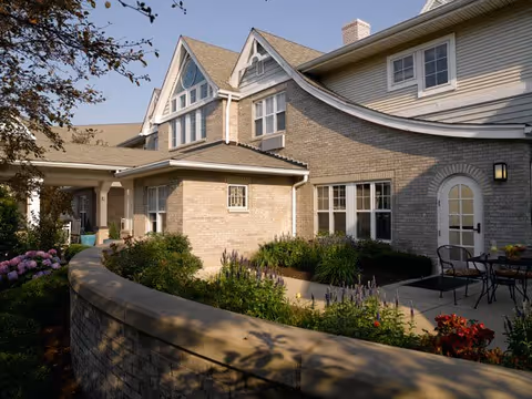 Exterior view of a brick senior living building with a landscaped courtyard, patio seating, and gabled rooflines.