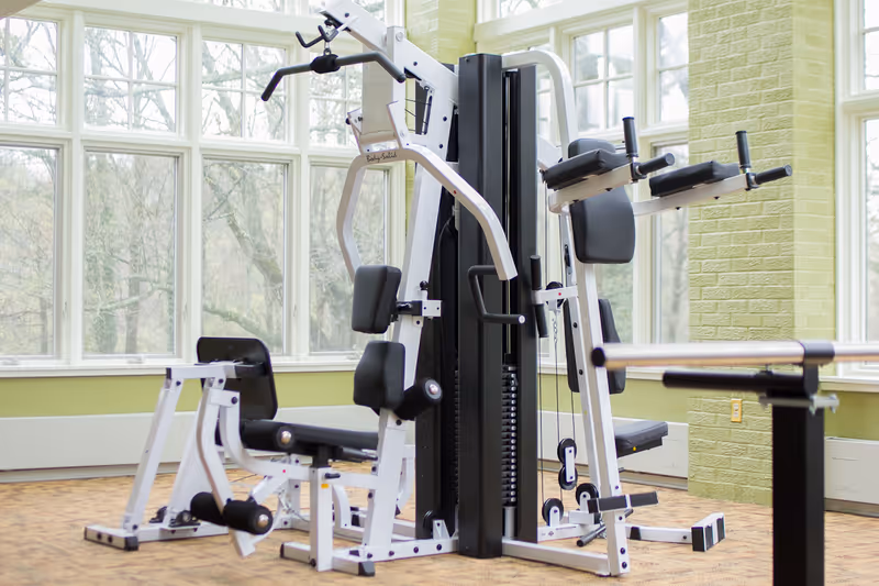 A multi-station gym machine in a bright room with large windows and green walls, designed for strength training exercises.
