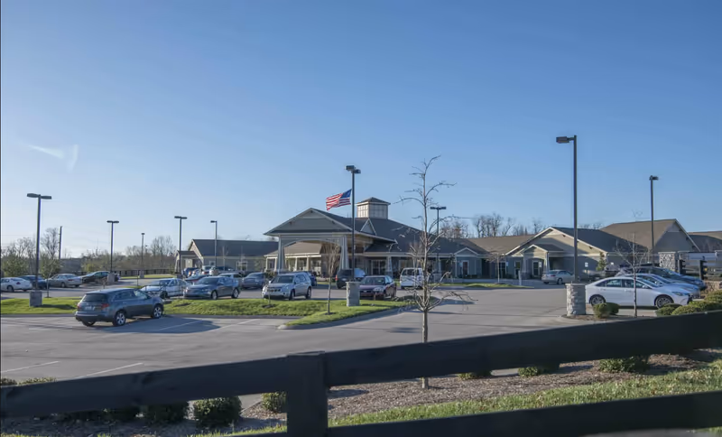 Front exterior view of a senior living building with a parking lot, several parked cars, and an American flag flying near the entrance.