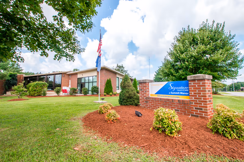 Exterior view of Signature HealthCARE at Summit Manor Rehab & Wellness Center showing a single-story brick building with large windows, a well-maintained lawn, landscaped bushes, and two flagpoles with flags. A brick sign with the facility's name is prominently displayed in the foreground.