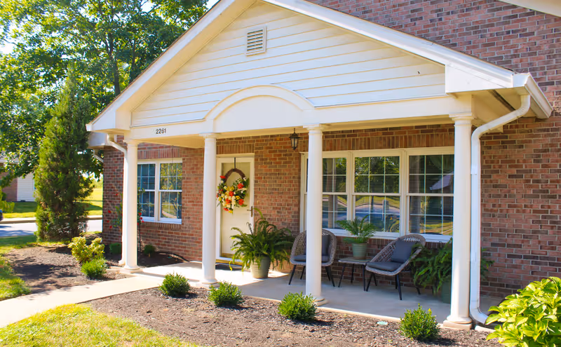 Front porch of a brick building with white columns and a white door decorated with a floral wreath. Two chairs and a small table with a potted plant are on the porch. There are small bushes and plants in the garden beds around the porch.