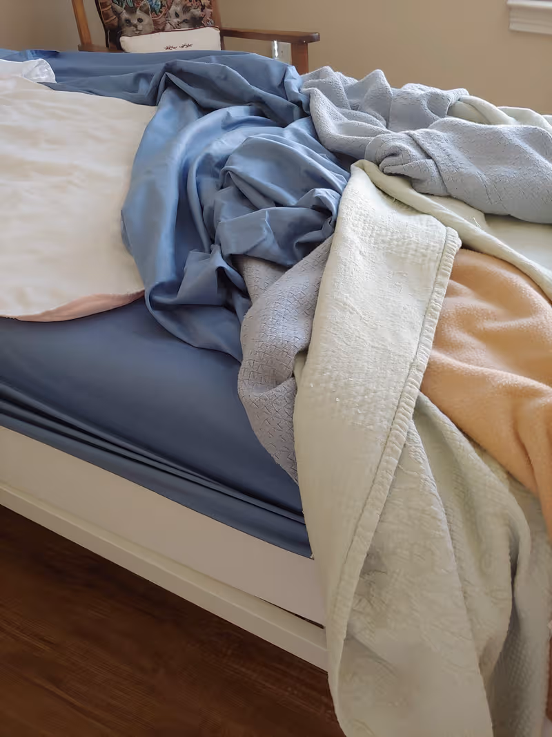 Unmade bed with blue sheets and blankets piled in a bedroom, chair with cat-themed cushion.
