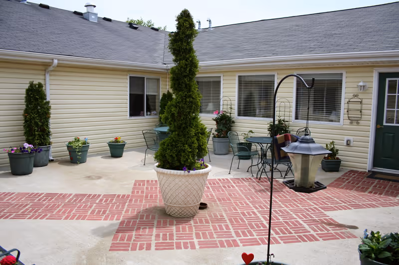A courtyard patio with potted plants, a large central planter, outdoor table and chairs, and a bird feeder in front of a single-story building.