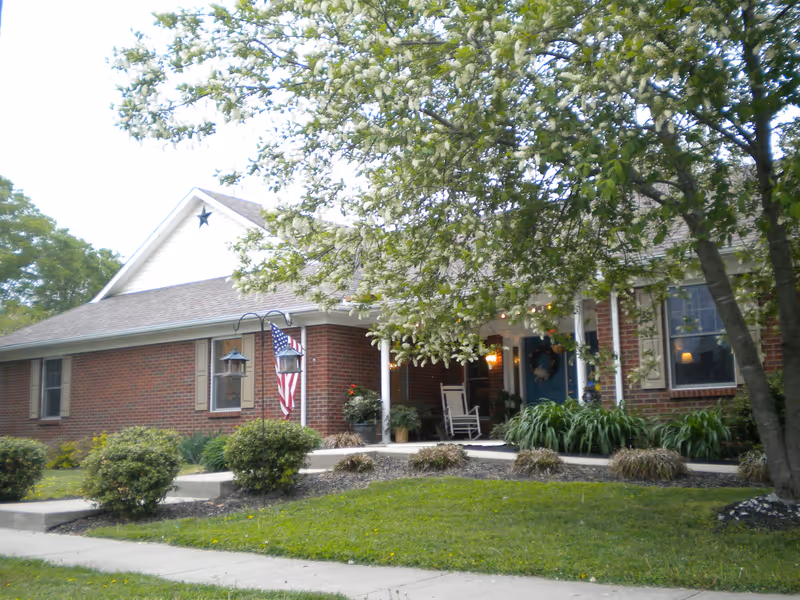 Brick house front with a covered porch featuring rocking chairs, an American flag, and a tree-shaded lawn.