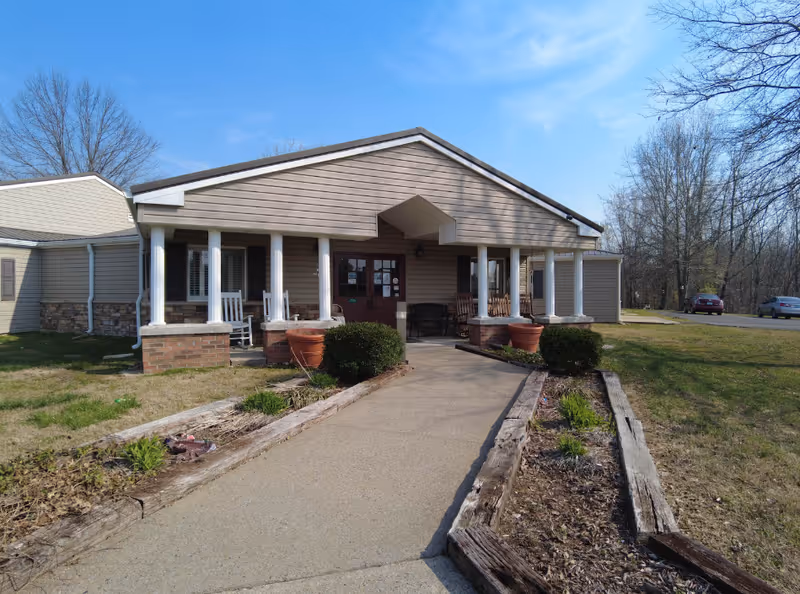Front exterior view of a single-story building with beige siding and a covered porch supported by white columns. There are rocking chairs on the porch and a concrete walkway leading to the entrance. The surrounding area has grass, some plants, and a few parked cars in the background under a clear blue sky.