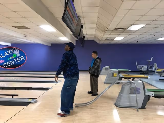 Two men standing in a bowling alley with multiple lanes. One man is preparing to bowl while the other watches. The bowling alley has a purple wall with a sign that reads 'Galaxy Center'. Overhead screens display scores above the lanes.