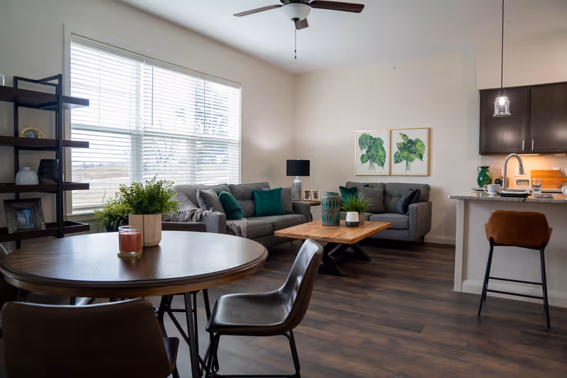 A cozy living room and dining area with a round wooden table and four chairs in the foreground. The living room has two gray sofas with green and gray cushions, a wooden coffee table with decorative plants, and two framed botanical prints on the wall. A ceiling fan is mounted above, and large windows with white blinds let in natural light. To the right, there is a kitchen counter with a brown bar stool and pendant lighting.