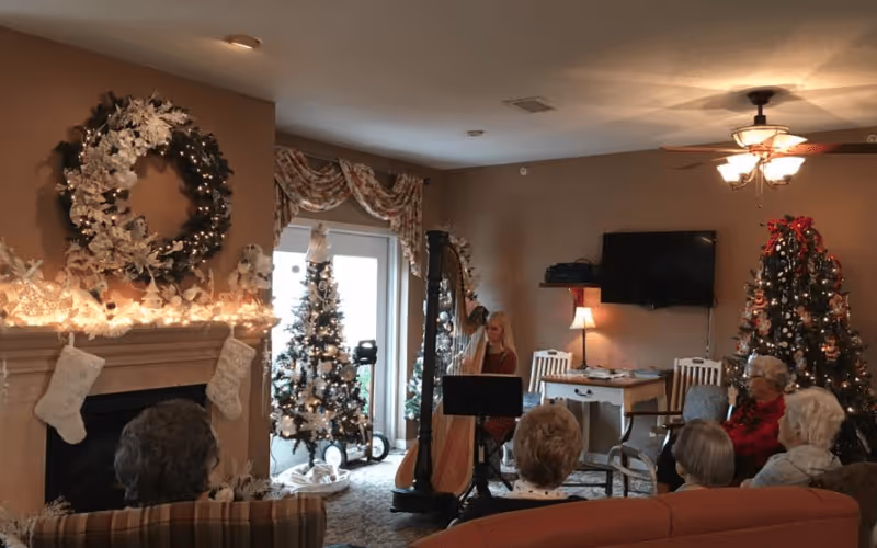 A cozy living room decorated for Christmas with a large wreath above the fireplace, two decorated Christmas trees, and stockings hanging on the mantle. Several elderly people are seated facing a woman playing a harp near a window with floral curtains. A ceiling fan with lights and a wall-mounted TV are visible in the background.