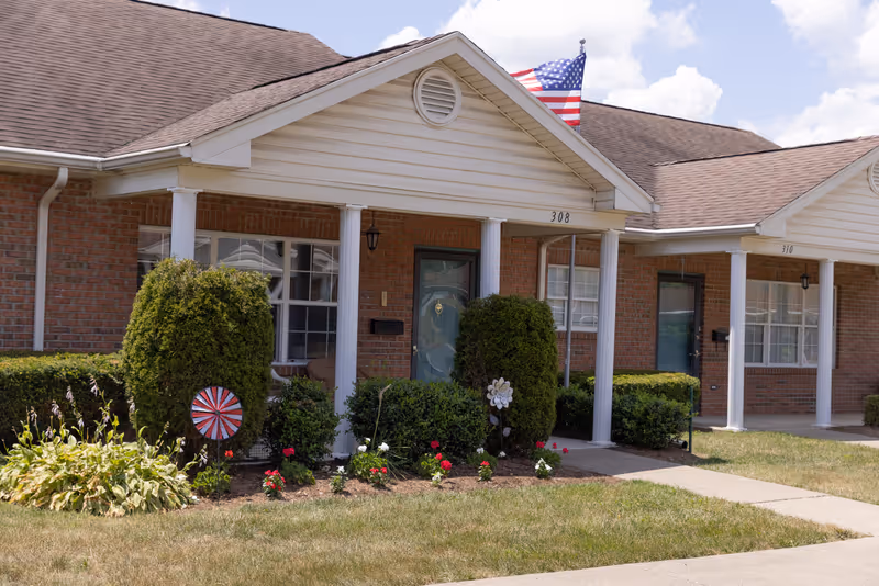 Brick senior living complex with front entrances, columns, and manicured landscaping under a blue sky