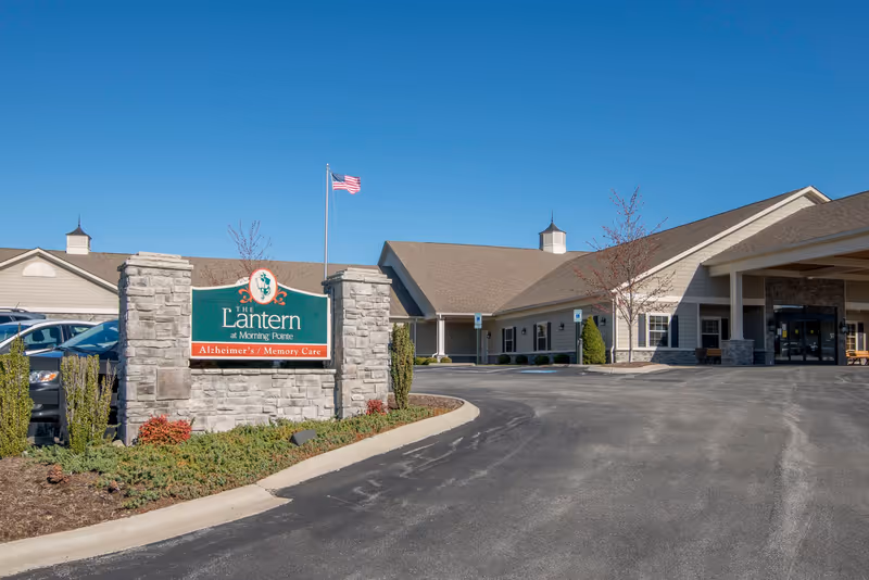 Front entrance of The Lantern at Morning Pointe Alzheimer's memory care building with a stone sign, driveway, and an American flag.