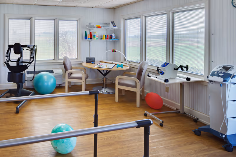 A bright rehabilitation/exercise room with parallel bars, exercise machines, therapy balls and a small table and chairs by large windows.