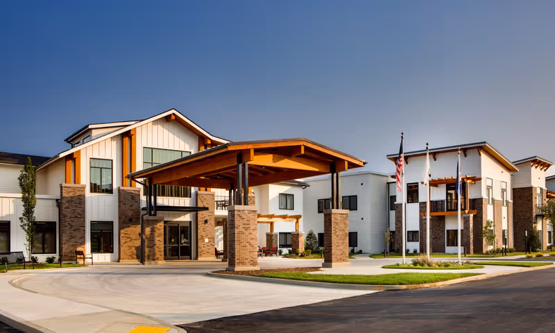 Exterior view of Preston Greens Senior Living facility showing a modern two-story building with white siding and brick accents, a covered entrance with wooden beams, three flagpoles with flags, and a paved driveway under a clear blue sky.