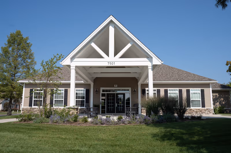 Front exterior view of a single-story building with beige siding, white trim, and a peaked roof. The entrance features a covered porch with white pillars and a triangular gable. There are several windows with black shutters on either side of the double glass doors. The building is surrounded by green grass, shrubs, and trees under a clear blue sky.